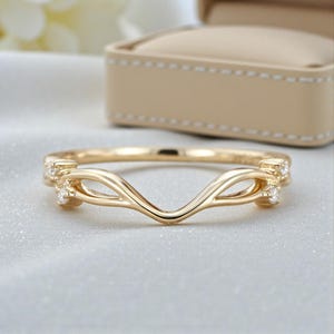 May include: A gold-colored ring with a curved design, featuring small, clear gemstones. The ring has an elegant, flowing shape with a delicate appearance. The ring is set against a soft, neutral background, suggesting a jewelry display.