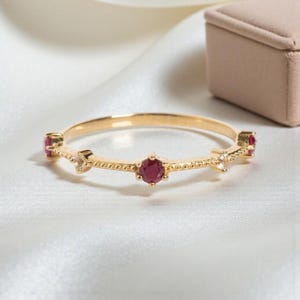 Puede incluir: Un delicado anillo de oro con una serie de pequeñas piedras preciosas redondas de color rojo intenso y piedras de acento transparentes. El anillo tiene una banda texturizada y está sobre un fondo blanco suave. Una pequeña caja de joyería beige está en el fondo.