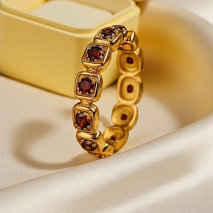 May include: A gold ring set with square-cut, deep red gemstones. The ring is shown on a white satin surface, with a yellow jewellery box in the background. The ring's design features a repeating pattern of square stones.
