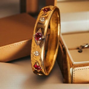 May include: A gold-coloured bangle bracelet featuring three red gemstones and two clear gemstones. The bracelet is displayed next to a gold-coloured jewellery box. The gemstones are set in a starburst pattern.