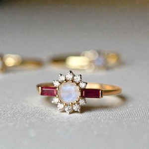 May include: A gold ring with a central moonstone surrounded by small diamonds. Two rectangular ruby stones flank the moonstone. The ring has a delicate, floral-like design, with other rings blurred in the background.