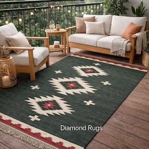 May include: Dark green outdoor rug with a geometric pattern in cream and red. The rug has a fringed edge and the text "Diamond Rugs" is visible. The rug is placed on a wooden deck, with outdoor furniture in the background.