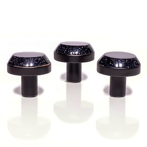 May include: Three black cabinet knobs with a mushroom design. Each knob features a dark blue top with sparkling flecks, edged with a gold band. The knobs are displayed on a white background.