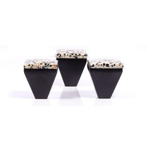 May include: Three black, geometric stools with speckled, cream-coloured tops. The stools have a trapezoidal shape, with the base wider than the top. The speckled pattern on the seat resembles a dalmatian's coat. The stools are set against a white background.