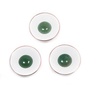 Puede incluir: Tres cuencos de cerámica blanca con borde color cobre y diseño de círculos concéntricos. Cada cuenco contiene una piedra verde lisa y redonda. Disposición sobre fondo blanco.