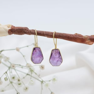 Pode incluir: Um par de brincos de ametista com molduras douradas. Os brincos estão pendurados em um galho marrom, com flores brancas e um fundo branco. As pedras de ametista têm uma forma irregular e facetada.
