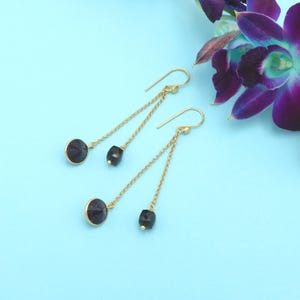 Może przedstawiać: Para złotych kolczyków wiszących. Każdy kolczyk ma okrągły, ciemnoniebieski kamień na górze, mały czarny kamień i złoty łańcuszek. Kolczyki są umieszczone na jasnoniebieskim tle z fioletowymi kwiatami.