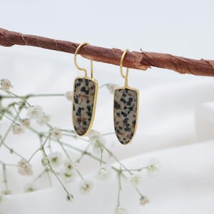May include: Gold-coloured earrings featuring a speckled, light-coloured stone set within a gold frame. The earrings are suspended from a brown branch, with white flowers and a white backdrop.