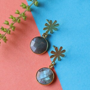 May include: A pair of gold-colored earrings with a flower-shaped stud and a round, faceted, gray gemstone pendant. The earrings are set against a pink and blue background, with a sprig of green leaves in the upper left corner.