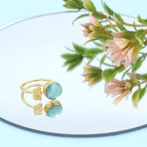 Pode incluir: Um anel dourado com uma pedra preciosa azul clara e dois pequenos detalhes de borboleta. O anel é exibido em uma superfície reflexiva com um fundo desfocado de elementos florais rosa e verdes. O anel é uma joia.