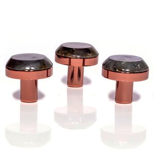 Puede incluir: Tres pomos de armario de color cobre con un diseño en forma de hongo. Cada pomo presenta una parte superior oscura y reflectante y un vástago cilíndrico. Los pomos están dispuestos en fila sobre un fondo blanco, con sus reflejos visibles.