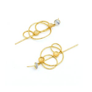 Può includere: Un paio di orecchini dorati dal design unico. Ogni orecchino presenta una serie di anelli interconnessi e sfere strutturate. Una piccola pietra di colore chiaro è incastonata all'estremità di ogni orecchino.