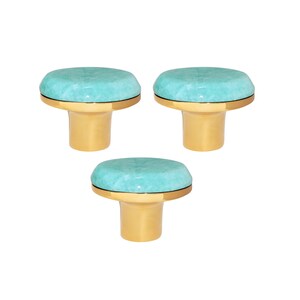 Op de afbeelding: Drie decoratieve kastknoppen. Elke knop heeft een ronde, lichtblauwe bovenkant en een goudkleurige basis. De knoppen hebben een gladde, gepolijste afwerking, tegen een witte achtergrond.