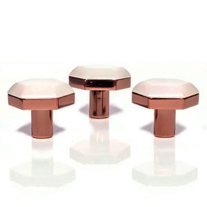 Pode incluir: Três puxadores decorativos para armários com um design geométrico. Cada puxador apresenta um topo octogonal branco com uma borda em ouro rosa e uma haste em ouro rosa a condizer. Os puxadores estão dispostos sobre uma superfície branca.