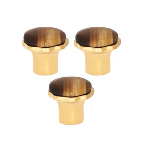 May include: Three cabinet knobs. Each has a gold-coloured base and a brown, oval-shaped stone top. The knobs have a decorative, scalloped edge where the stone meets the base. The knobs are set against a white background.
