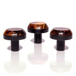 Puede incluir: Tres tiradores decorativos para armarios. Cada tirador presenta una base y un vástago negros, coronados por una cúpula facetada de color ámbar y marrón, similar al vidrio. Los tiradores están dispuestos en fila sobre un fondo blanco, con sus reflejos visibles debajo.
