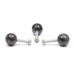 Può includere: Tre pomelli decorativi per mobili. Ogni pomello presenta una pietra sferica scura e maculata attaccata a un bullone color argento con un dado esagonale. I pomelli sono disposti su uno sfondo bianco.