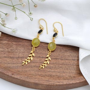 May include: A pair of gold-coloured drop earrings. Each earring has a black gemstone, a light green oval gemstone, and a gold-coloured leaf design. The earrings are displayed on a wooden surface with white fabric and small white flowers.
