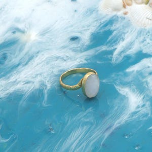 May include: A gold ring featuring a large, oval, white gemstone. The ring's band has a textured pattern. The ring is set against a blue and white background, with seashells in the top right corner.