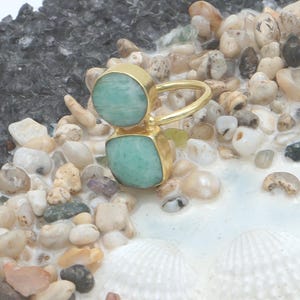 May include: A gold ring with two light green gemstones, one round and one square. The ring is displayed on a bed of pebbles and seashells. The gemstones have a marbled effect. The ring's design evokes a coastal theme.