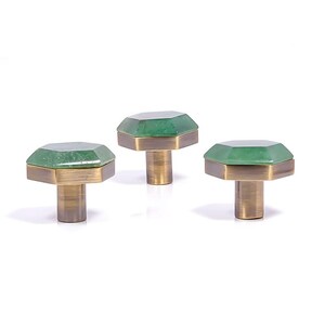 May include: Three cabinet knobs with green, faceted tops and brass-coloured bases. The knobs have an octagonal shape. The knobs are set against a white background. These knobs are ideal for adding a touch of elegance to furniture.
