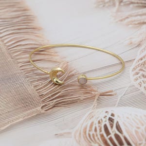 Puede incluir: Pulsera de puño abierta de color dorado con una luna creciente y una pequeña piedra redonda de color rosa claro. La pulsera es una pieza de joyería delicada, perfecta para el uso diario. El diseño es sencillo y elegante.