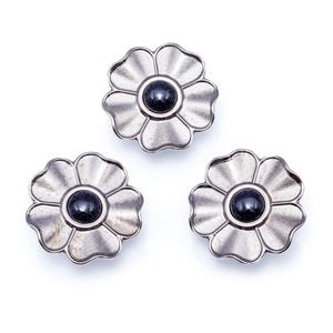 Könnte beinhalten: Drei silberfarbene, blütenförmige Knöpfe mit einem dunklen, möglicherweise schwarzen Mittelstein. Die Blütenblätter haben eine geriffelte Textur, und die Knöpfe sind auf weißem Hintergrund angeordnet. Diese dekorativen Knöpfe könnten für Kleidung oder Bastelprojekte verwendet werden.