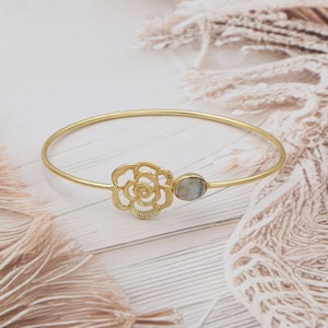 May include: Gold-toned bangle bracelet featuring a rose-shaped filigree design and a small, oval-shaped stone. The bracelet is set against a light-colored wooden surface with soft, neutral-toned fabric in the background.