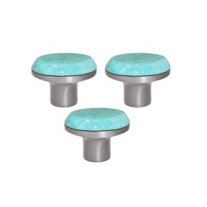 Può includere: Tre pomelli per armadi con una parte superiore maculata color turchese chiaro e una base argentata spazzolata. I pomelli hanno una forma arrotondata, a forma di fungo, e sono posti su uno sfondo bianco. Questi pomelli sono ideali per aggiungere un tocco di colore a qualsiasi stanza.