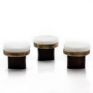 May include: Three decorative cabinet knobs. Each knob features a white, circular top, a brass-colored band, and a dark brown, rectangular base. The knobs are arranged in a row against a white background.