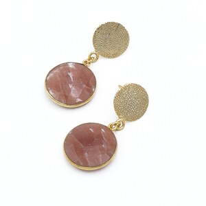 May include: A pair of gold-toned earrings. Each earring features a textured, circular stud and a larger, round, faceted, pink gemstone pendant. The gemstones are set in gold-colored bezels. The earrings are connected by a small gold loop.
