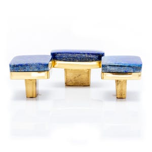 Può includere: Tre pomelli per mobili con piani quadrati in lapislazzuli blu intenso e basi in ottone color oro. I pomelli sono disposti in fila, mostrando il loro design elegante e la loro maestria artigianale. Ogni pomello ha una forma di base diversa.