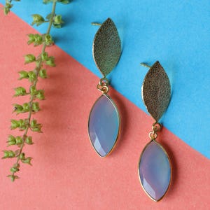May include: A pair of gold-colored earrings with a leaf-shaped top and a light blue, oval-shaped gemstone pendant. The earrings are set against a backdrop of pink and blue.