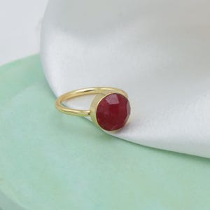 May include: A gold ring featuring a large, faceted, oval-shaped ruby gemstone. The ring's band is a simple, polished gold. The ruby is a deep, rich red color, and the ring is set against a white fabric background.