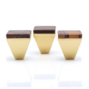 Puede incluir: Tres tiradores decorativos para armarios. Cada tirador presenta una parte superior cuadrada de ojo de tigre y una base triangular dorada. Los tiradores están dispuestos en fila sobre un fondo blanco, con sus reflejos debajo.