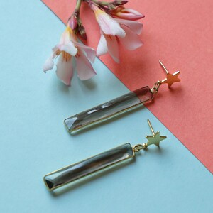 May include: Gold-coloured earrings featuring a rectangular, smoky-grey crystal pendant and a small star detail. The earrings are set against a blue and coral backdrop, with pink flowers in the upper left corner.