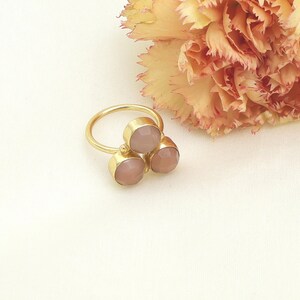 Può includere: Un anello dorato con tre pietre preziose rotonde rosa chiaro. L'anello è posto su uno sfondo bianco morbido, con un fiore color pesca nell'angolo in alto a destra. Le pietre sono incastonate in castoni d'oro individuali.