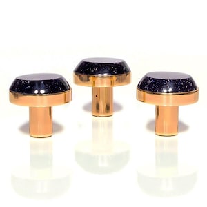 Pode incluir: Três rolhas de garrafa douradas com um topo azul escuro salpicado. As rolhas têm uma haste cilíndrica e uma base larga e plana. Os topos têm uma faixa dourada metálica. As rolhas estão dispostas em fila contra um fundo branco.