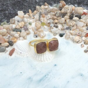 Op de afbeelding: Een gouden ring met twee vierkante, roodbruine edelstenen. De ring ligt op een witte zeeschelp, tegen een achtergrond van kleine kiezelstenen en een lichtblauwe en witte ondergrond.
