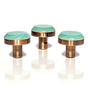 Könnte beinhalten: Drei dekorative Schrankknöpfe mit mintgrüner Oberseite und Messingbasis. Die Knöpfe haben eine abgerundete Form und sind vor weißem Hintergrund in einer Reihe angeordnet. Die Knöpfe werden wahrscheinlich für Möbel oder Schränke verwendet.