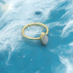 Può includere: Un anello dorato con una fascia strutturata e una pietra a forma di pera di colore chiaro. L'anello è presentato su uno sfondo blu e bianco, che mette in risalto il design del gioiello.