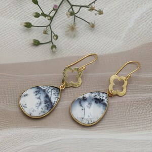 May include: A pair of gold-colored dangle earrings. Each earring features a teardrop-shaped, white stone with black and gray veining, set in a gold-colored frame. The top of each earring has a gold-colored clover-shaped design.