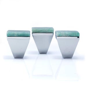 May include: Three silver rings with a geometric design. Each ring features a rectangular, light green stone set atop a triangular silver band. The rings are displayed against a white background, showcasing their modern aesthetic.