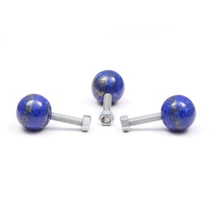May include: Three blue lapis lazuli sphere cabinet knobs with silver-colored bolts and nuts. The knobs are a deep blue with flecks of gold, and are arranged against a white background.