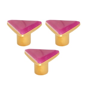 Può includere: Tre pomelli decorativi per mobili. Ogni pomello presenta una parte superiore triangolare magenta e una base dorata. Questi pomelli sono progettati per mobili e misurano circa 3 cm di altezza, aggiungendo un tocco di stile.