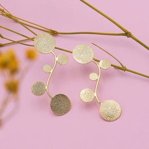 May include: Gold-coloured earrings with a botanical design. Each earring has a curved stem with textured, circular elements of varying sizes. The earrings are set against a pink background with dried plant sprigs.