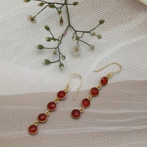 Può includere: Un paio di orecchini pendenti color oro con cinque pietre preziose rosse rotonde ciascuno. Gli orecchini hanno un semplice design a gancio e sono esposti su una superficie neutra e strutturata con delicati accenti floreali.