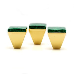 May include: Three gold rings with a geometric design. Each ring features a square, green, marbled stone set atop a gold band. The bands have a trapezoid shape, wider at the top and tapering down.
