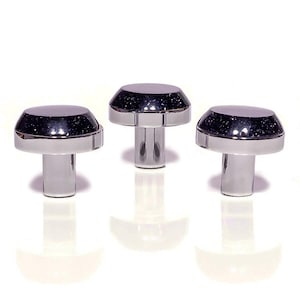 May include: Three chrome cabinet knobs with a mushroom-shaped design. The top of each knob is a dark, speckled colour, and the base is a shiny silver. The knobs are displayed against a white background.