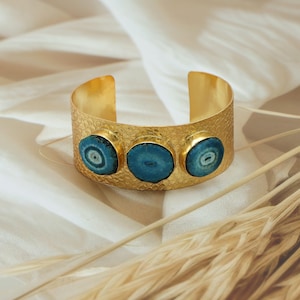 Może przedstawiać: Złota bransoleta na nadgarstek o teksturowanej powierzchni i trzech okrągłych, niebiesko-zielonych kamieniach. Otwarta bransoleta jest prezentowana na białej tkaninie.